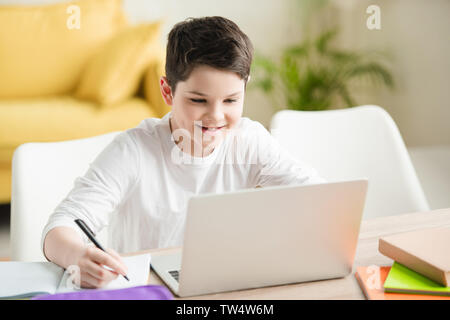 cheerful boy using laptop and writing in copy book while doing schoolwork at home Stock Photo
