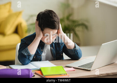 tired boy holding hands near head while sitting at table with notebooks and laptop Stock Photo