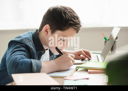 adorable attentive boy writing in notebook and using laptop while doing homework Stock Photo
