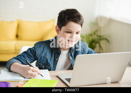 cheerful boy writing in notebook and using laptop while doing schoolwork at home Stock Photo
