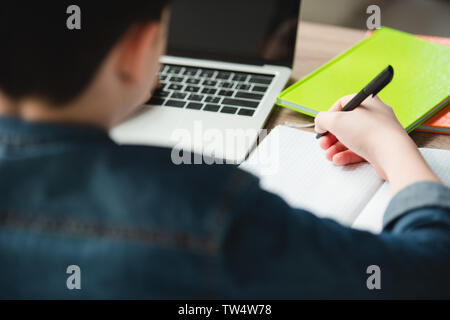 back view of boy writing in notebook and using laptop while doing homework Stock Photo