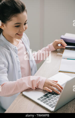 cheerful schoolchild using laptop while sitting at wooden desk and doing homework Stock Photo