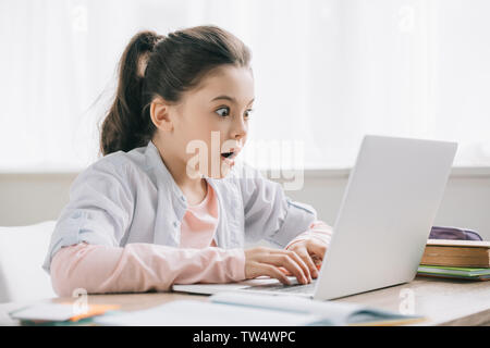 surprised child using laptop while sitting at desk and doing schoolwork at home Stock Photo