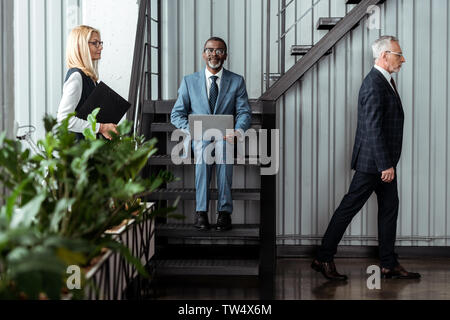 selective focus of handsome african american man in glasses using laptop while sitting on stairs near partners Stock Photo