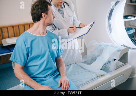 partial view of young radiologist showing diagnosis to patient sitting on ct scanner bed Stock Photo