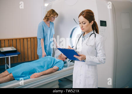 nurse preparing patient for mri scanning while radiologist writing on clipboard Stock Photo