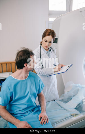 attentive doctor writing on clipboard while standing near patient sitting on ct scanner bed Stock Photo