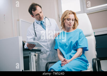 attentive doctor touching shoulder of upset woman while holding digital tablet Stock Photo