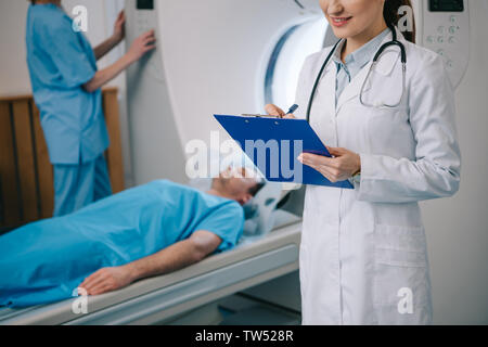 cropped view of smiling radiographer writing on clipboard while assistant standing near lying patient and operating ct scanner Stock Photo