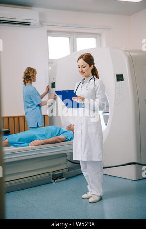 smiling radiographer writing on clipboard while assistant standing near lying patient and operating ct scanner Stock Photo
