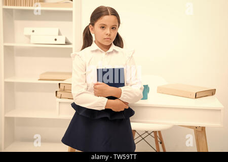 How make education more interesting for first formers. Girl child hold book while stand table white interior. Kid school uniform calm sad indifferent face hold book. Indifferent about knowledge. Stock Photo