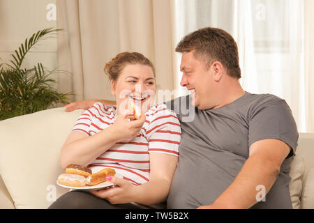 Overweight couple eating sweets on sofa at home Stock Photo