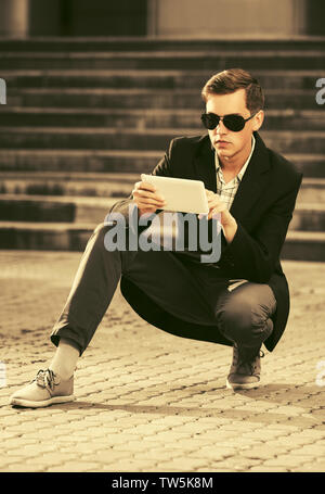 Young handsome business man using tablet computer on city street Stylish trendy male model wearing sunglasses and black suit jacket Stock Photo