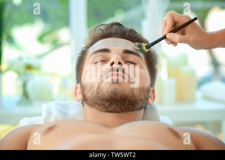 Beautician applying scrub onto young man's face in spa salon Stock Photo