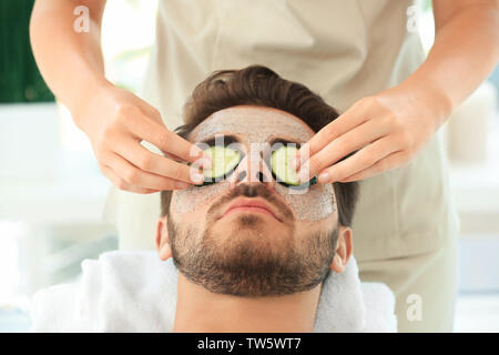 Beautician putting fresh cucumber slices on young man's face in spa salon Stock Photo