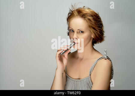 Portrait of young attractive beautiful red-haired woman with electronic cigarette on gray background in studio Stock Photo