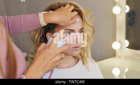 Stylish attractive blonde in beauty salon. Girl makes an evening hairstyle at professional hairdresser. Hair stylist uses spray-shaped hairspray to fi Stock Photo