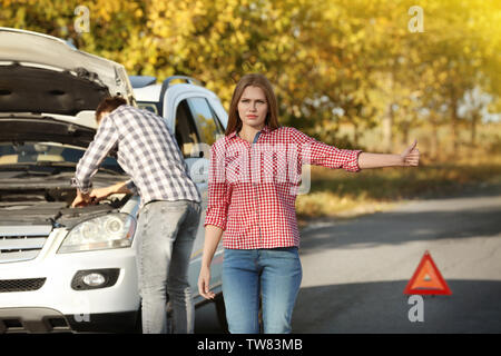 Young woman hitchhiking on road while man trying to repair broken car Stock Photo
