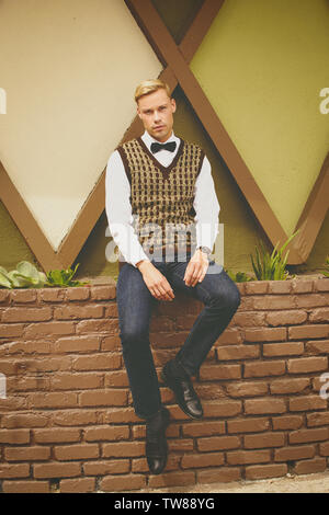 White Caucasian Male sits on a brick wall edge, wearing a vest, bow tie and jeans. He looks relaxed. A handsome male model. Stock Photo