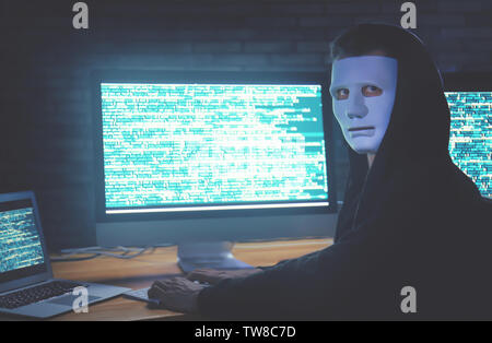 Masked hacker using computer in dark room. Threat of cyber attack Stock Photo