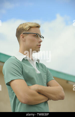 A Caucasian white male wearing a green vintage 60s polo shirt and vintage eyeglasses,,arms folded looking away. A blue sky background behind him. Stock Photo