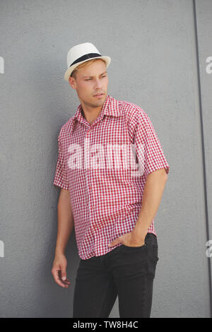 A blonde male model wearing a red western plaid shirt, white fedora hat, posing against a wall standing outdoor, looking away. Stock Photo