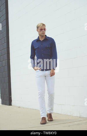 A Caucasian Blonde male model posing in a vintage 70s blue shirt, posing outdoor against a white wall background. Stock Photo