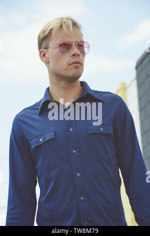 A Caucasian Blonde male model posing in a vintage 70s blue shirt, posing outdoor against a blue sky background. A vintage fashion editorial. Stock Photo