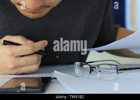 man taking notes from smartphone device. writing and reading. glasses place on desk in front of a pile of papers. man holding pen in office. business Stock Photo