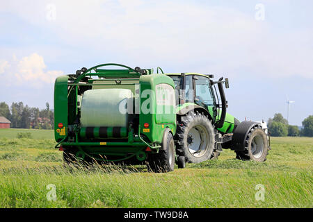 Salo, Finland. June 15, 2019. Deutz-Fahr tractor and McHale 3 plus baler baling silage in green plastic sheet in hay field on a sunny day of summer. Stock Photo