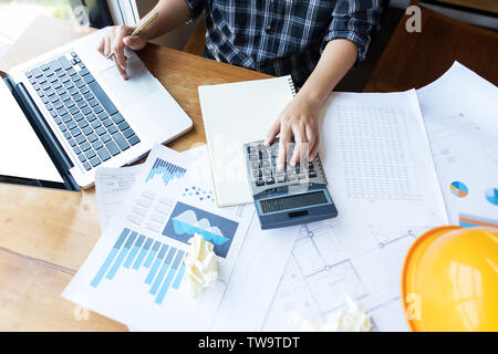 Asian engineer , architect woman using calculator for accounting and analyzing building structure from blueprint and paper chart in front of computer Stock Photo