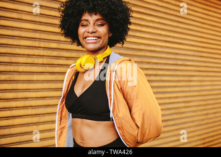 Sporty young woman standing outdoors in the city and smiling. Female athlete with headphones resting after workout. Stock Photo