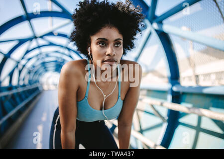 Young female athlete leaning over and looking away. Motivated and focused sporty woman before a run on walkway bridge. Stock Photo