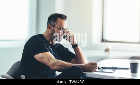 Entrepreneur looking at his digital tablet and talking on cell phone while sitting at his desk. Businessman busy working in office. Stock Photo