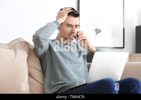 Dissatisfied young man talking on phone while working with laptop at home Stock Photo