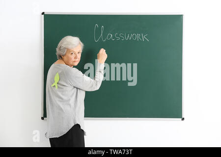 Senior teacher with paper fish on her back writing on chalkboard. April fool's day celebration Stock Photo