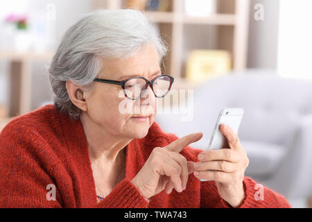 Mature woman with hearing aid using smartphone indoors Stock Photo