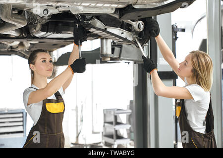 Two female mechanics standing under car in workshop and repairing chassis of auto. Strong women in coveralls and protective gloves working in auto salon and fixing vehicles. Concept of maintenance. Stock Photo
