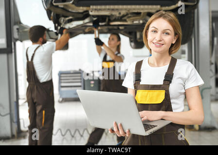 Front view of attractive woman working at auto service. Professional engineer in coverall keeping computer, looking at camera and smiling while colleagues repairing car. Concept of maintenance. Stock Photo