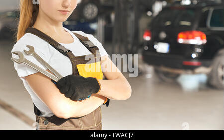 Serious woman in white shirt, coverall and protective gloves posing in workshop. Skilled female mechanic with folded arms holding wrench in auto service. Concept of repairing and fixing cars. Stock Photo