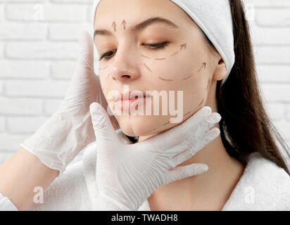 Closeup of attractive woman sitting in beauty salon before lifting procedure. Young lady with markup on face looking down and smiling while hands of doctor looking at skin. Concept of plastic surgery. Stock Photo