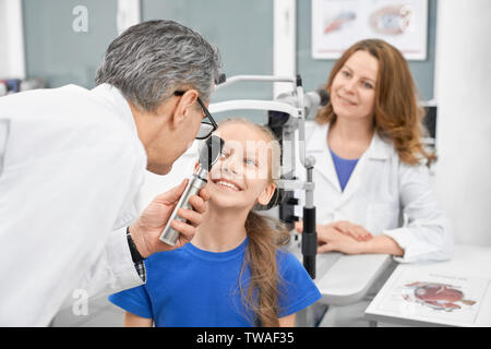Girl checking vision in laboratory. Child sitting and looking at camera while professional male oculist testing sight with special tool. Eyes doctors working in optical store. Concept of medicine. Stock Photo