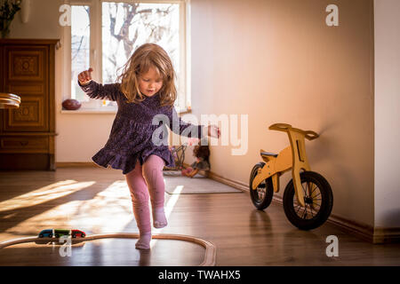 Happy, little child girl playing and dancing inside toy train tracks, in a sunlit room with window, having fun during her playtime