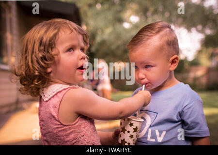 Little adorable girl generously sharing her drink with her cute friend, letting him taste it, from a birthday cup with stars, using a straw Stock Photo