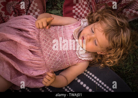 Little beautiful, adorable girl, with curly hair, in cute, pink retro dress, relaxing outside, laying on blankets, on grass Stock Photo