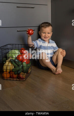 Little kid sitting on kitchen floor, picking fresh red tomato out of vegetable basket