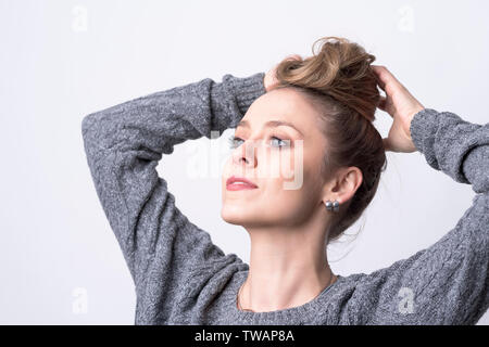 Portrait of independent beautiful young woman making hair bun hairdo herself on light gray background. Stock Photo