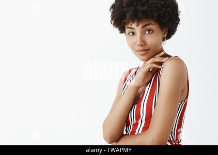 Waist-up shot of good-looking flirty and feminine dark-skinned woman with curly hairstyle touching shoulder and crossing chest with hand smiling Stock Photo