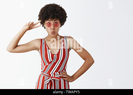 Studio shot of good-looking silly and playful dark-skinned girl with curly hairstyle, holding hand on hip, showing peace sign and folding lips Stock Photo