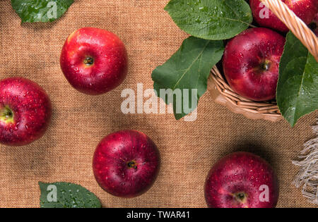 Close up of fresh wet red apples and green leaves covered by water drops in small basket and on sacking material. Selective focus. Healthy eating conc Stock Photo